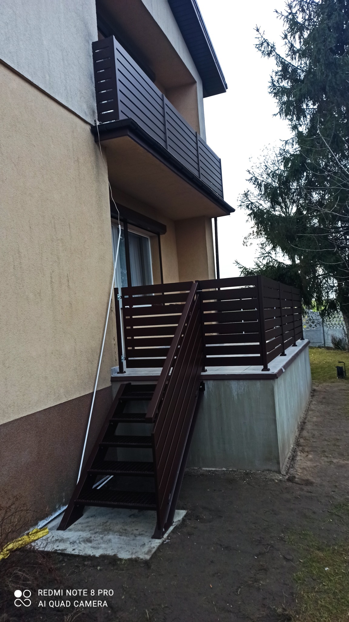 Zewnętrzne schody z brązową, metalową konstrukcją i ażurową balustradą prowadzące na balkon z podobną balustradą, widoczne z poziomu gruntu przy budynku.