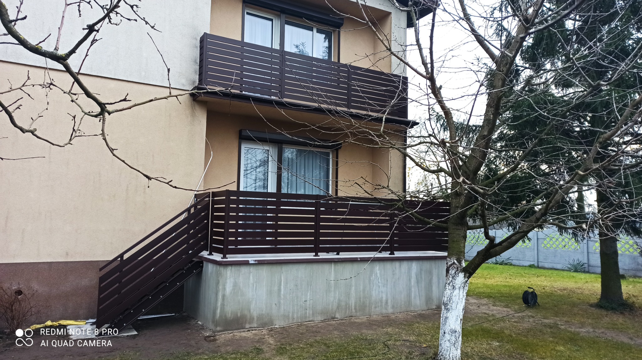 Brązowe, poziome panele tworzące balustradę balkonu i ogrodzenie schodów zewnętrznych, widoczne z poziomu gruntu, drzewo z bielonym pniem na pierwszym planie.