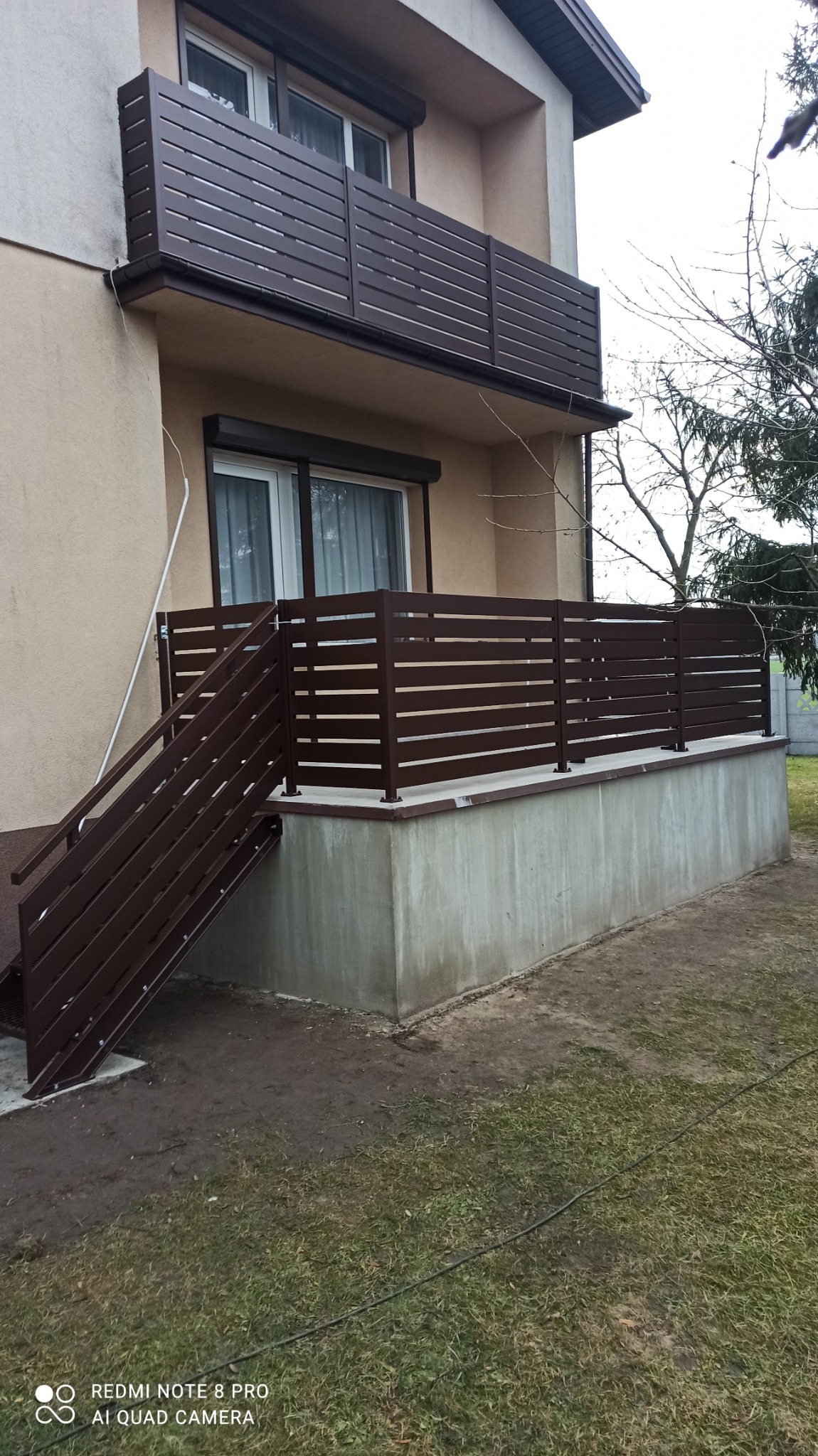 Nowoczesne, poziome, brązowe panele tworzące balustradę balkonu i ogrodzenie tarasu, z dopasowanymi kolorystycznie schodami, na tle beżowego budynku z białymi oknami.