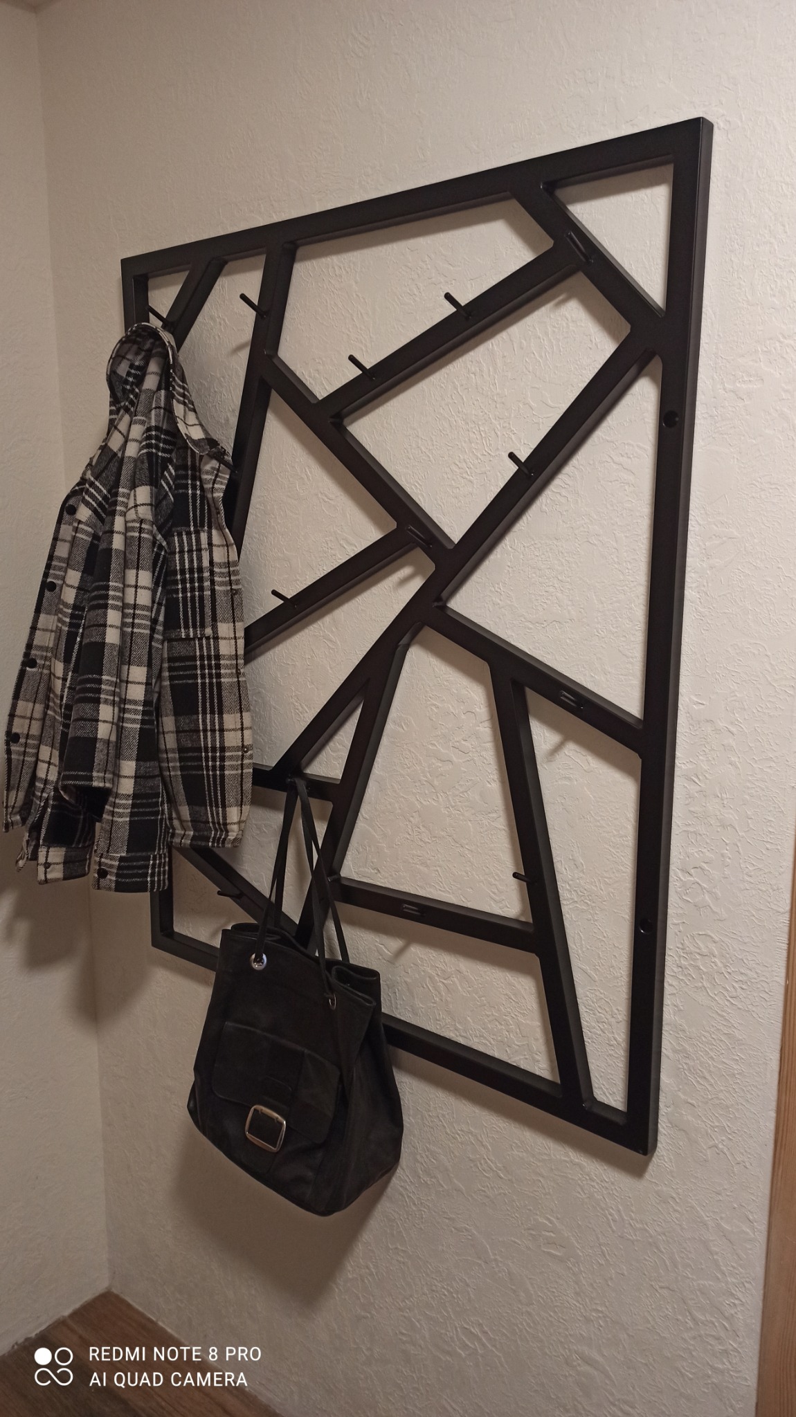 Czarny, geometryczny wieszak metalowy przymocowany do ściany, z zawieszoną koszulą w kratę i czarną torbą.