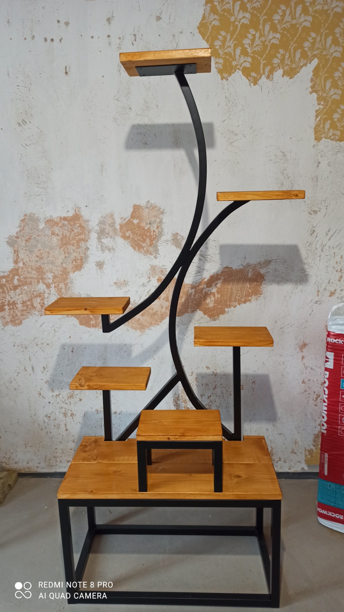 Wielopoziomowy kwietnik z drewnianymi półkami i czarną, giętą konstrukcją z metalu, na tle ściany z odpadającą tapetą.