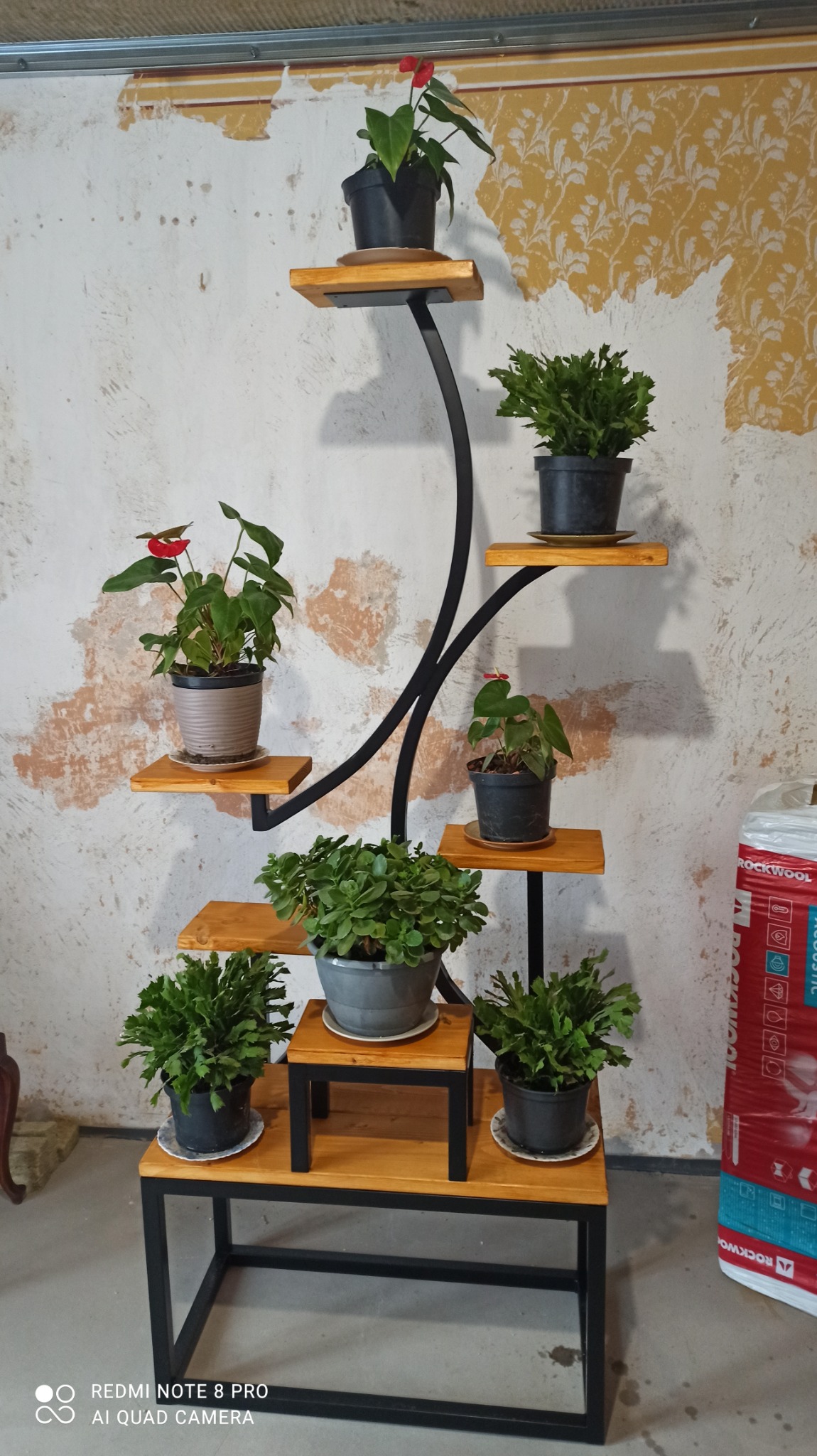 Wielopoziomowy kwietnik z metalową konstrukcją i drewnianymi półkami, na których ustawione są doniczki z różnymi roślinami, na tle ściany z widocznymi śladami tapety.