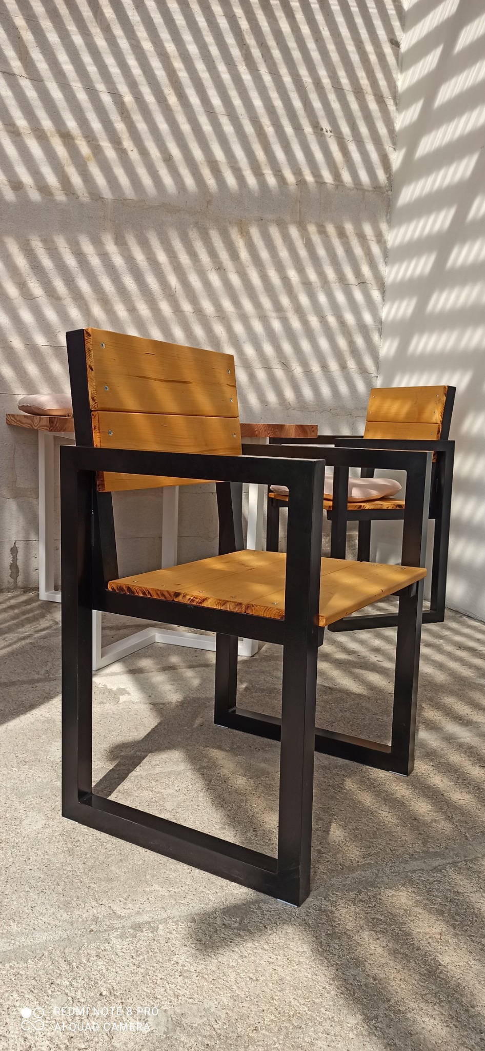 Dwa krzesła z drewnianymi siedziskami i oparciami na metalowych, czarnych ramach, ustawione na zewnątrz w cieniu rzucanym przez żaluzje.