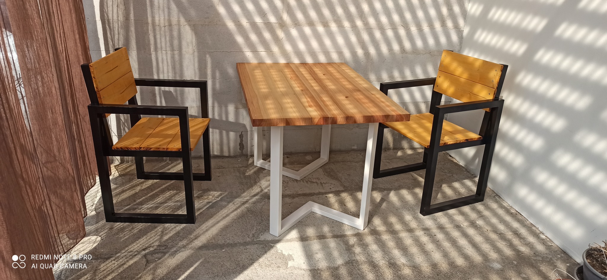 Stół z blatem z jasnego drewna i białymi nogami w stylu loftowym, otoczony dwoma krzesłami z czarnymi metalowymi ramami i drewnianymi siedziskami w ciepłym odcieniu.