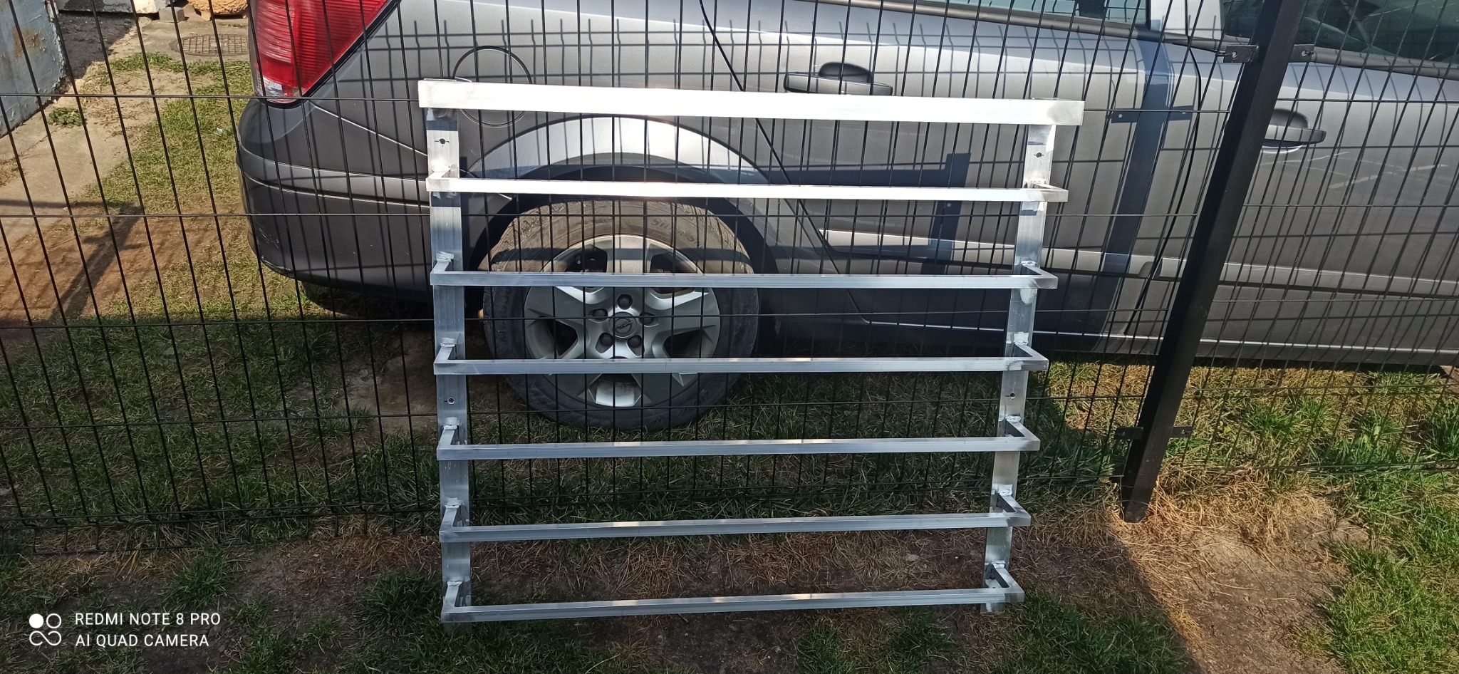 Surowa, aluminiowa konstrukcja na buty, z widocznymi spawami i otworami montażowymi, oparta o czarne ogrodzenie panelowe na tle szarego samochodu.