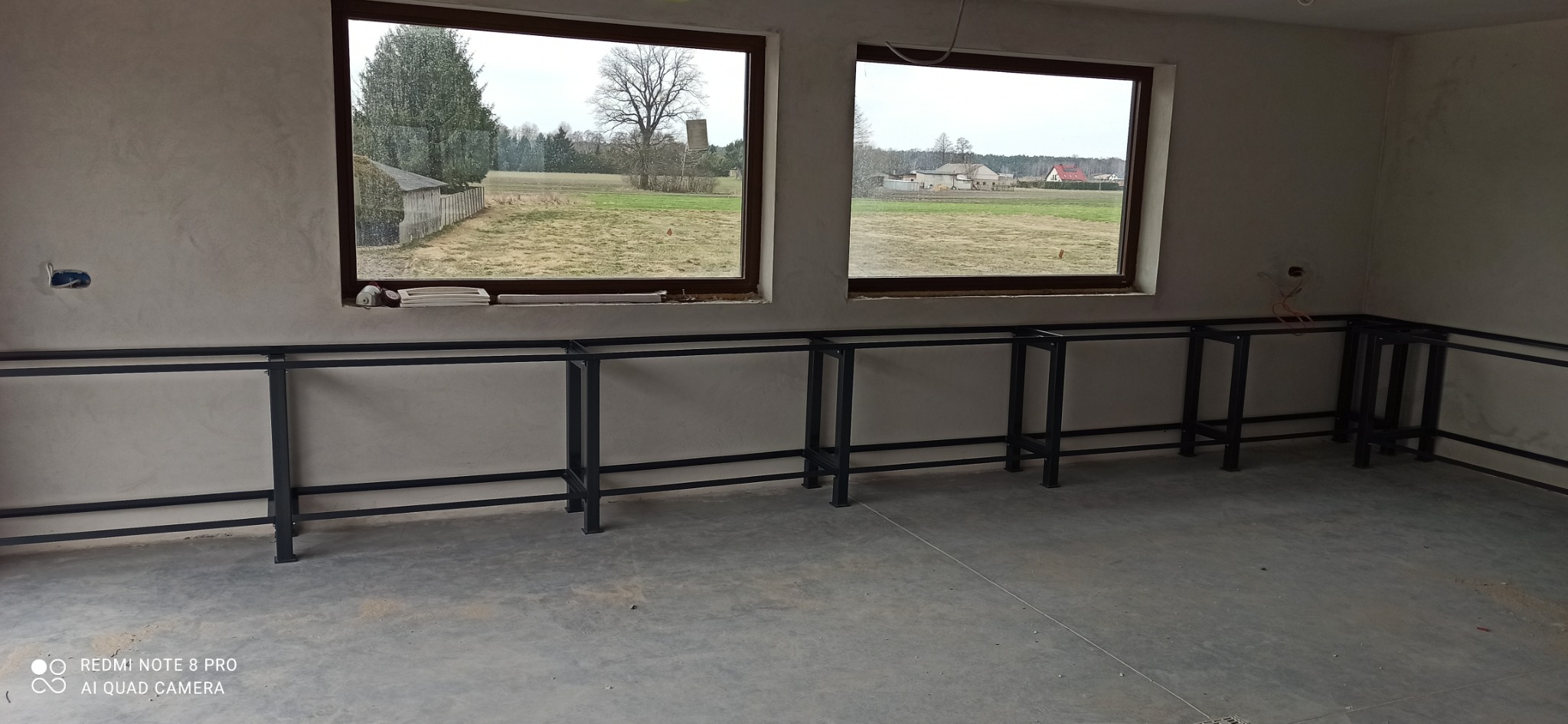 Metalowa konstrukcja pod zabudowę kuchenną w surowym wnętrzu z dwoma oknami, przez które widać wiejski krajobraz. Elementy instalacji elektrycznej na ścianie.