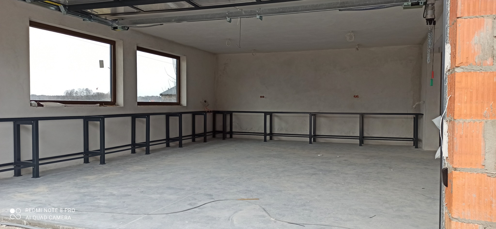 Wnętrze w stanie surowym z zamontowaną metalową konstrukcją pod blaty robocze wzdłuż ścian. Widoczne okna i instalacje elektryczne.