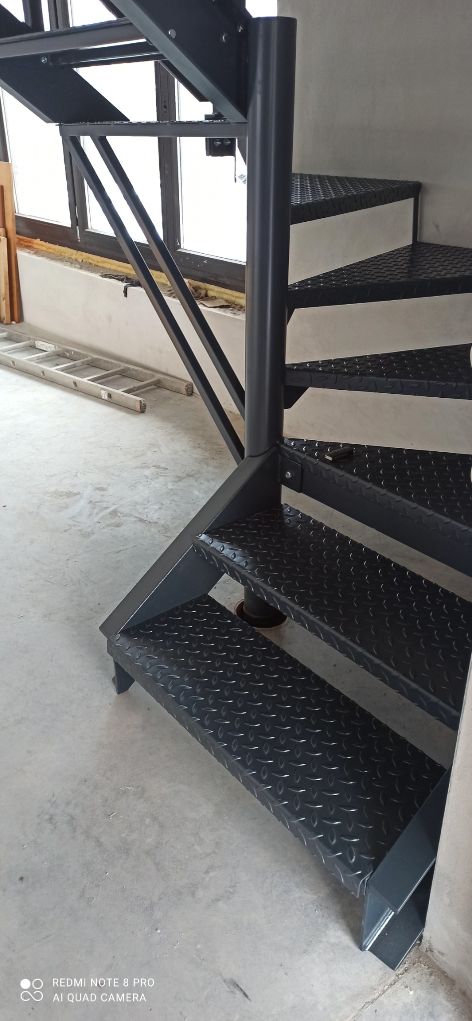 Ciemne, spiralne schody metalowe z blachy ryflowanej w surowym wnętrzu. W tle okno i drabina. Ujęcie z dołu, ukazujące konstrukcję i fakturę stopni.