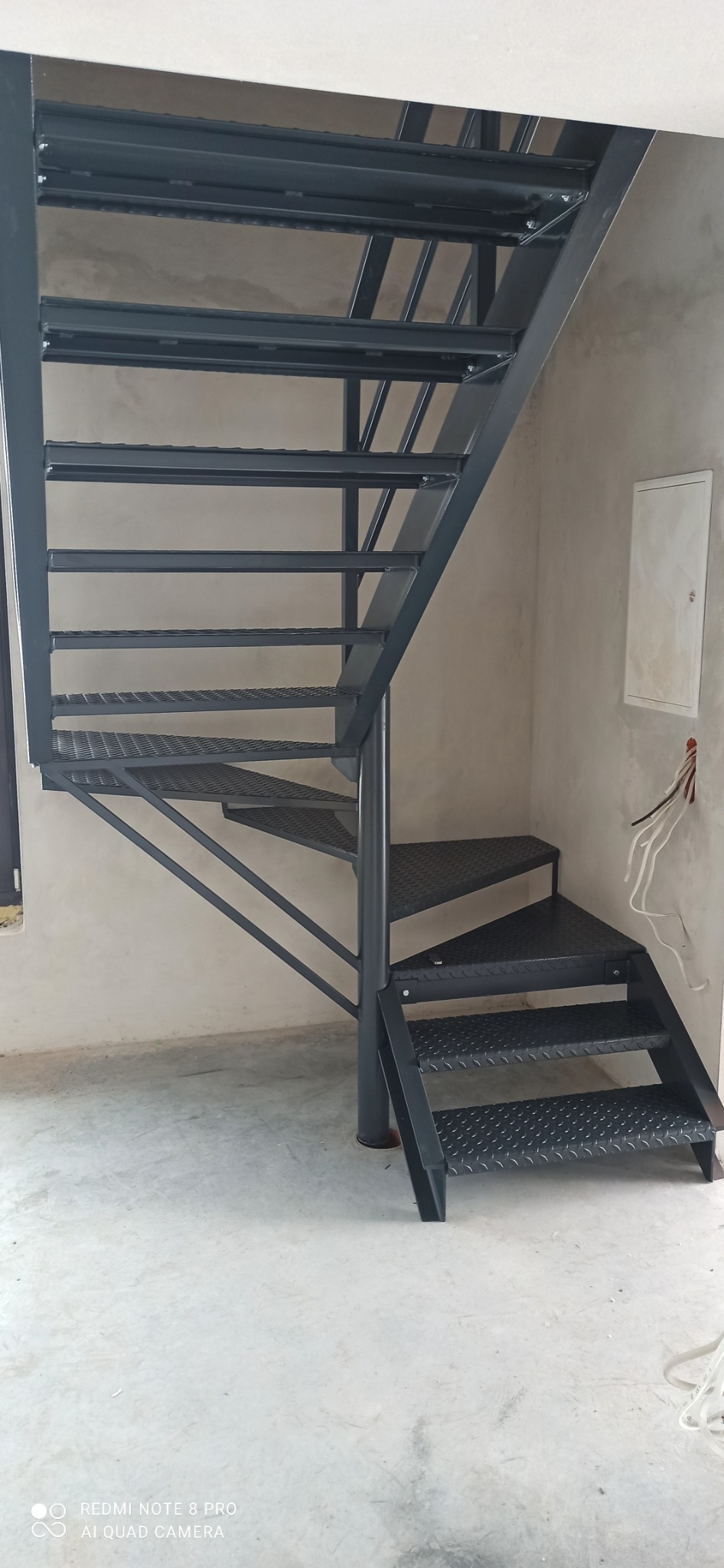 Metalowe schody spiralne w surowym wnętrzu, z widocznymi przewodami elektrycznymi i skrzynką na ścianie. Schody pomalowane na ciemny kolor, z perforowanymi stopniami.