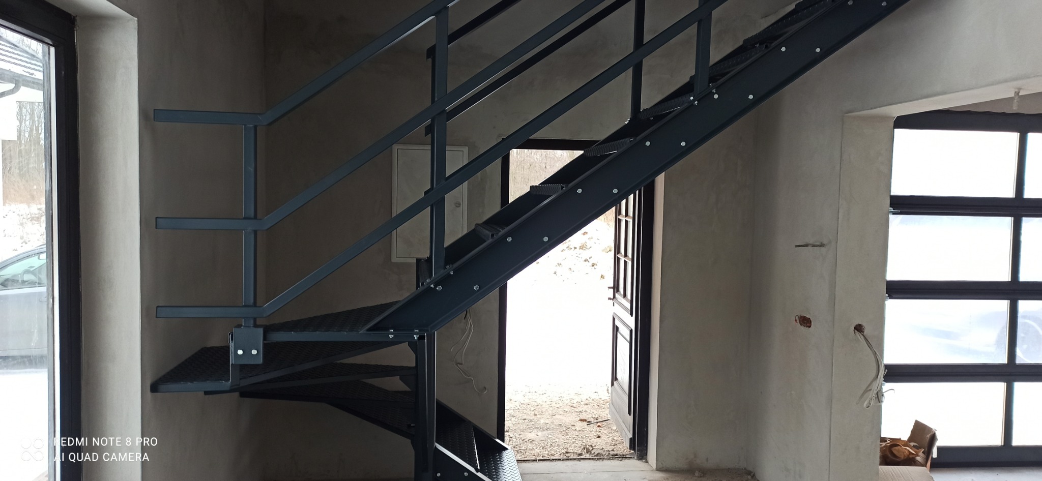 Nowoczesne, metalowe schody z ażurowymi stopniami w surowym wnętrzu. Widoczne detale mocowań i spawów. W tle okno i otwarte drzwi.