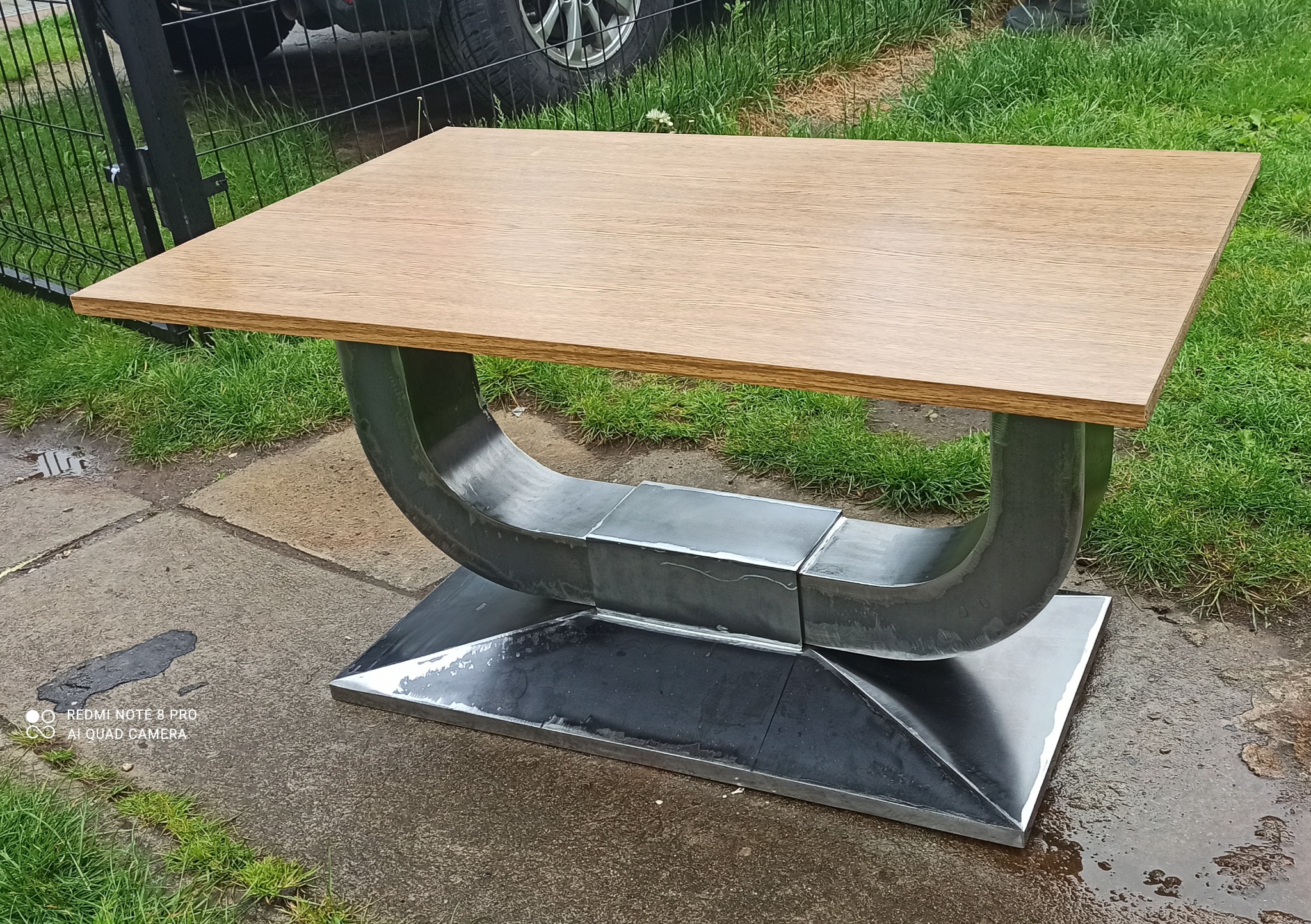 Stół z drewnianym blatem i metalową podstawą w kształcie litery U, spawana z elementów, stoi na zewnątrz na betonowej nawierzchni obok trawy. Widoczne ślady spawania.