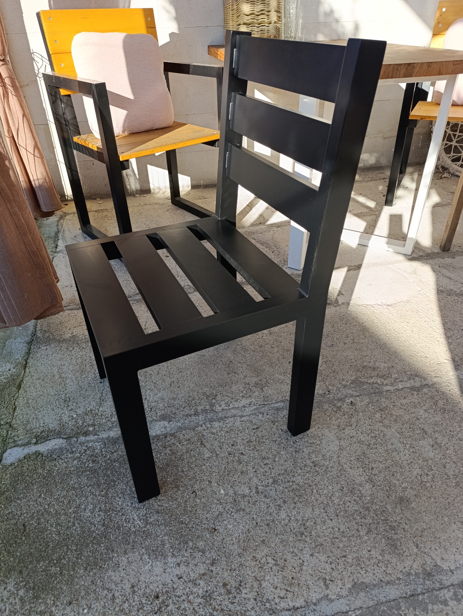 Nowoczesne, czarne krzesło metalowe z poziomymi szczeblami na oparciu, stojące na betonowej posadzce w słoneczny dzień. Widoczne inne meble w tle.