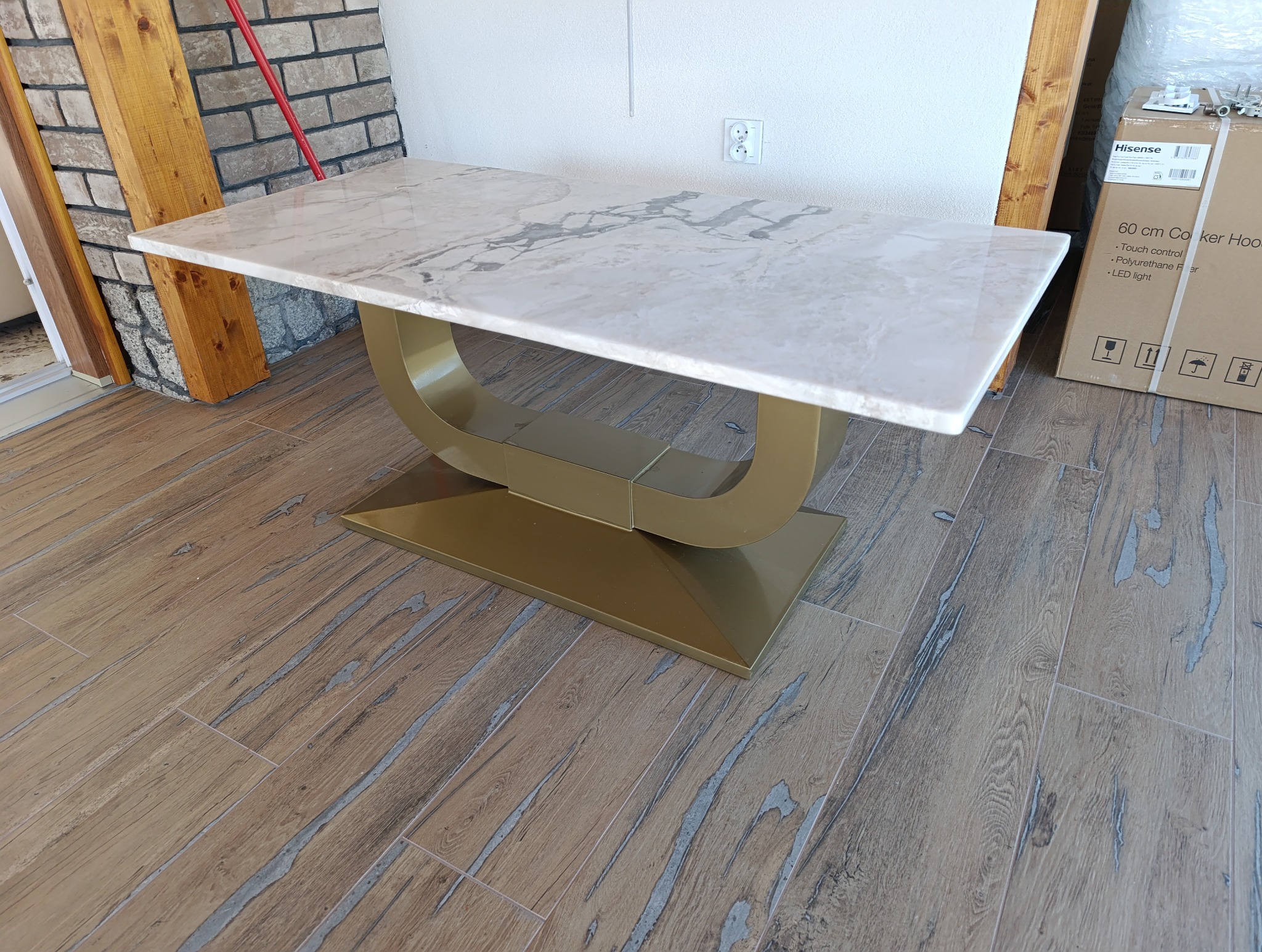 Stół z marmurowym blatem i złotą, metalową podstawą w kształcie łuku. Nowoczesny design, widoczne płytki podłogowe imitujące drewno. Częściowo widoczny karton.