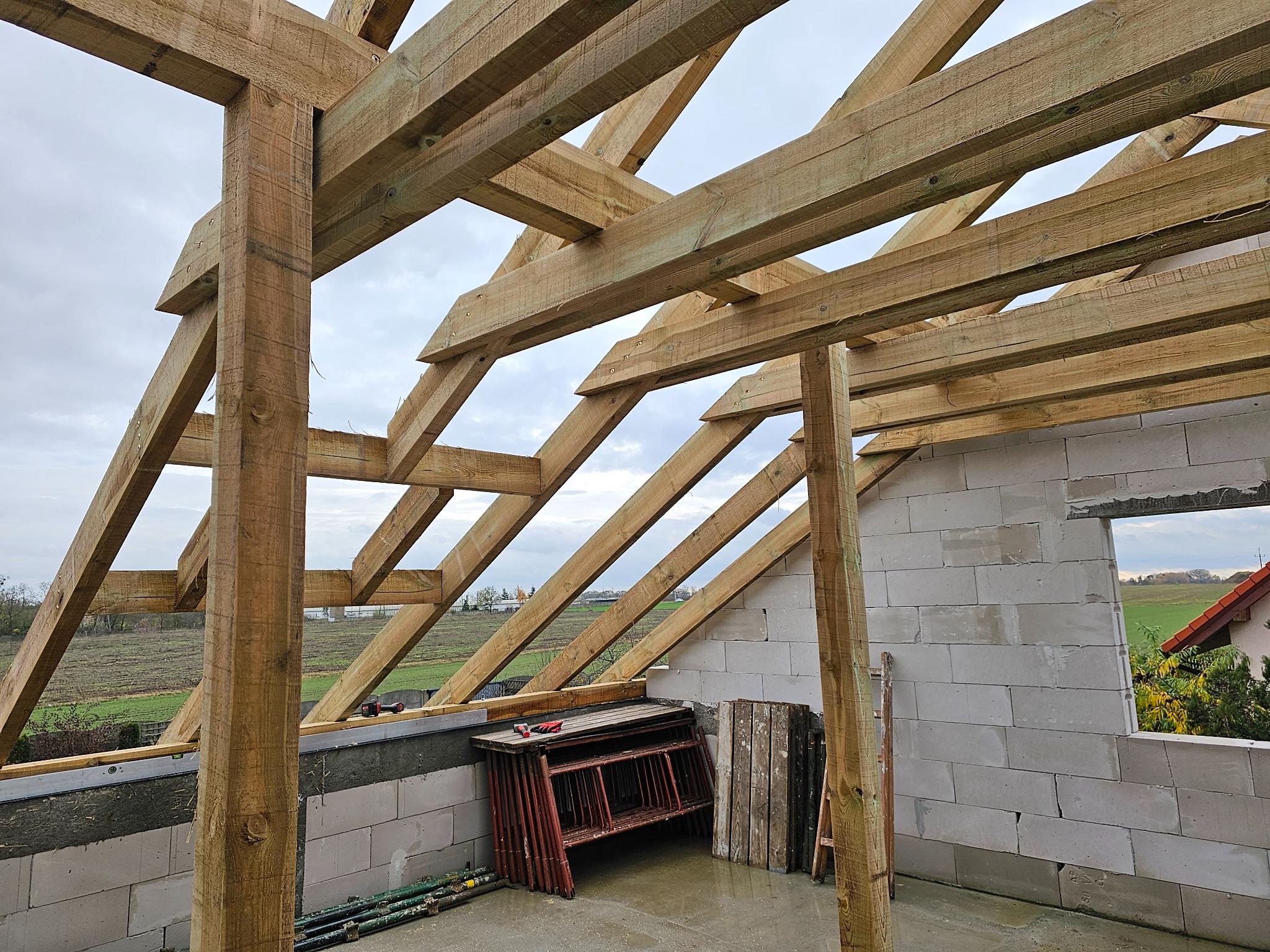 Surowa drewniana konstrukcja dachu w trakcie budowy, widok z wnętrza budynku, elementy więźby dachowej z widocznymi śrubami, ściany z bloczków betonowych, w tle krajobraz wiejski.
