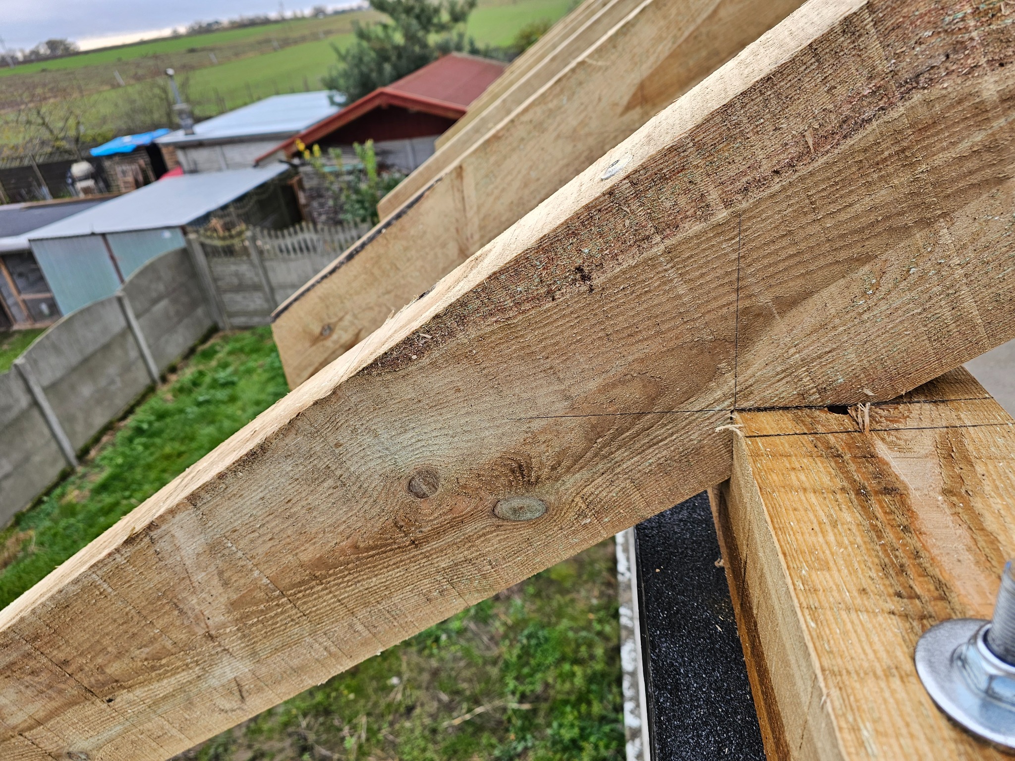 Wiązar dachowy z drewna, widoczne łączenia i elementy mocujące, w tle zabudowania w Buku.