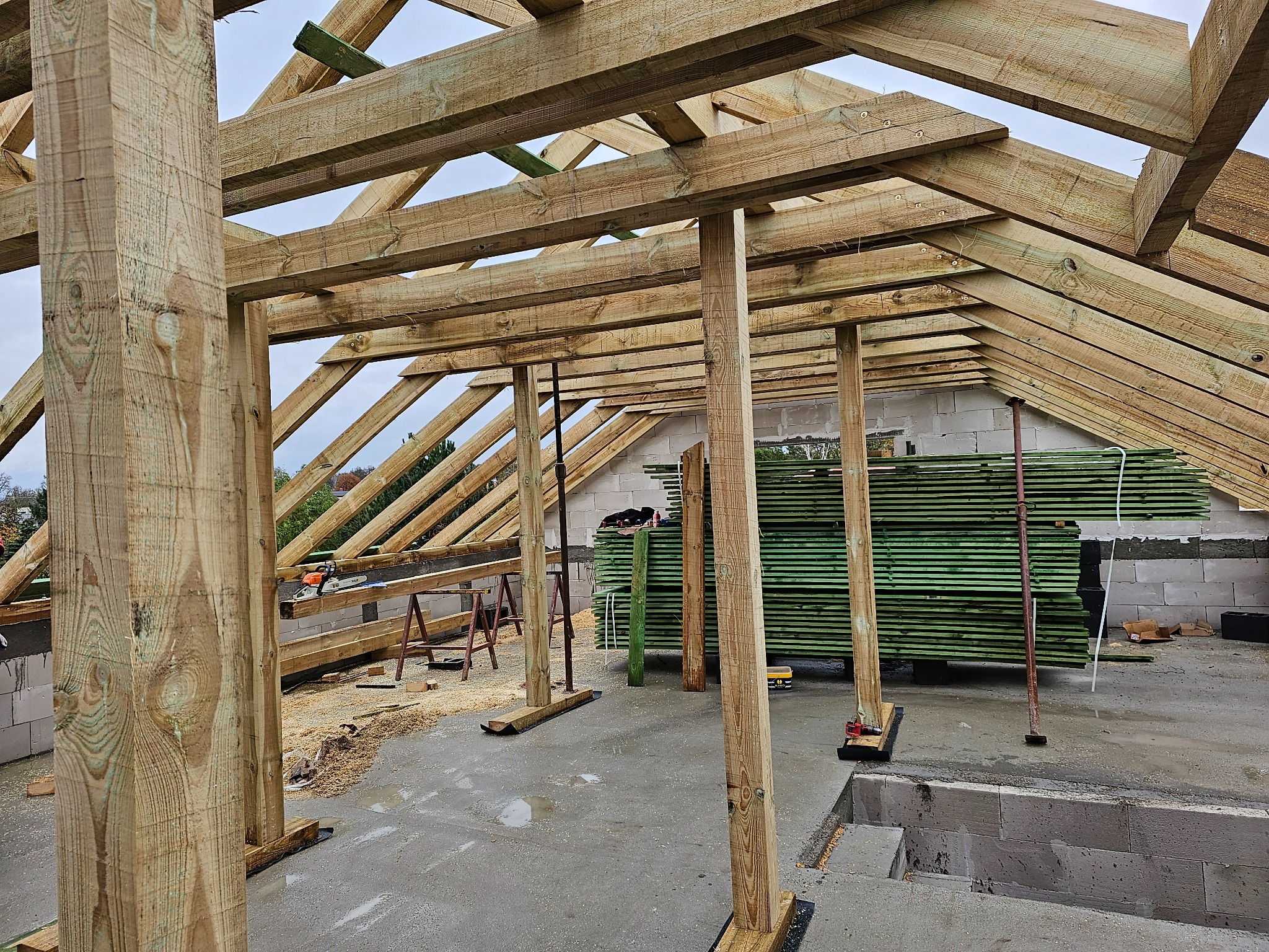 Wznoszenie drewnianej konstrukcji dachu z widocznymi słupami nośnymi, deskami i stertą zielonych łat, piła łańcuchowa leży na belce, surowy stan budowy.