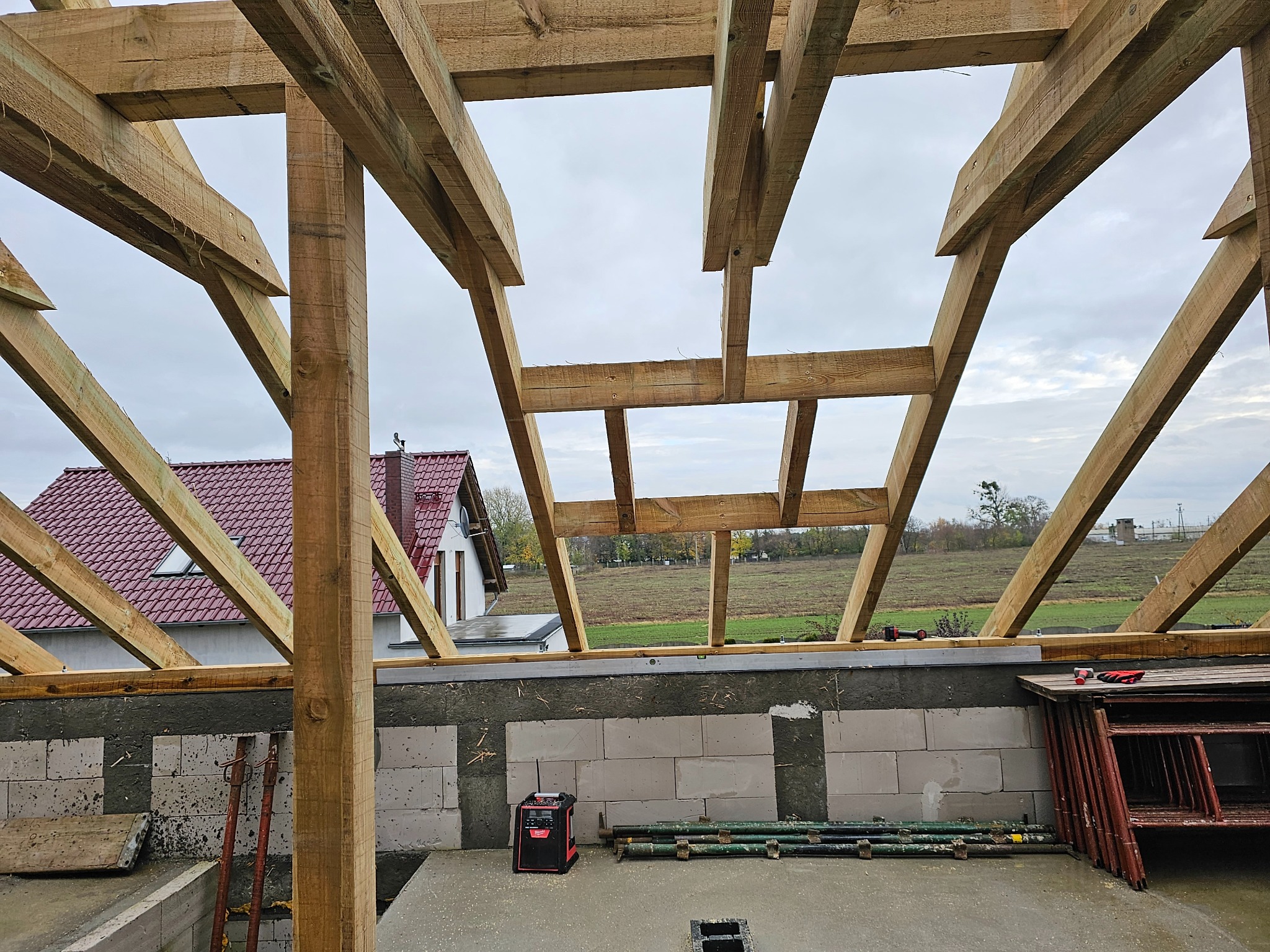 Drewniana konstrukcja dachu w trakcie budowy, widok z wnętrza, w tle dom z czerwonym dachem i krajobraz.