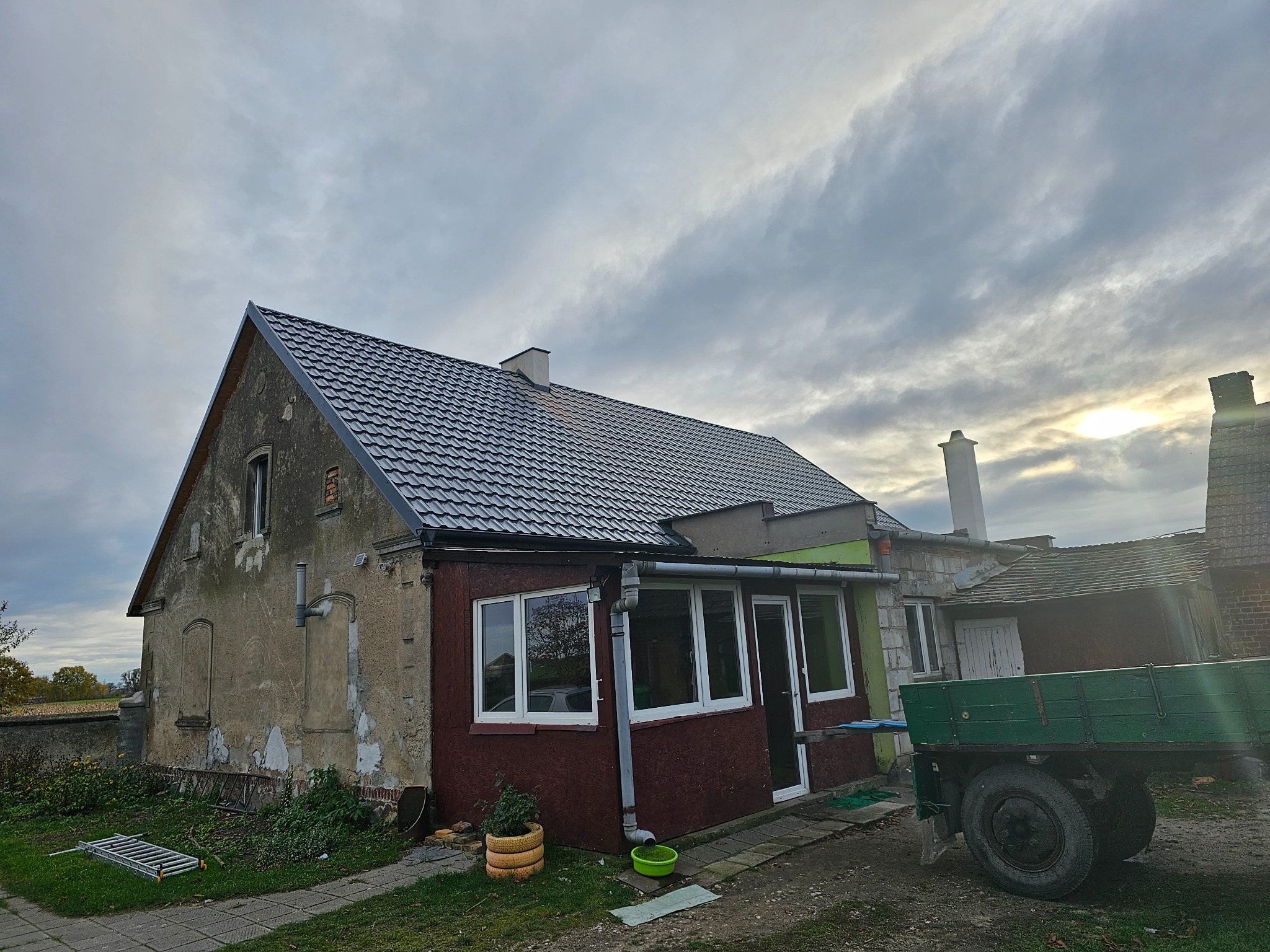 Dom z nowym, grafitowym pokryciem dachowym z blachy trapezowej, widoczny z boku, z elementami starej elewacji i podwórka z zielonym wozem.