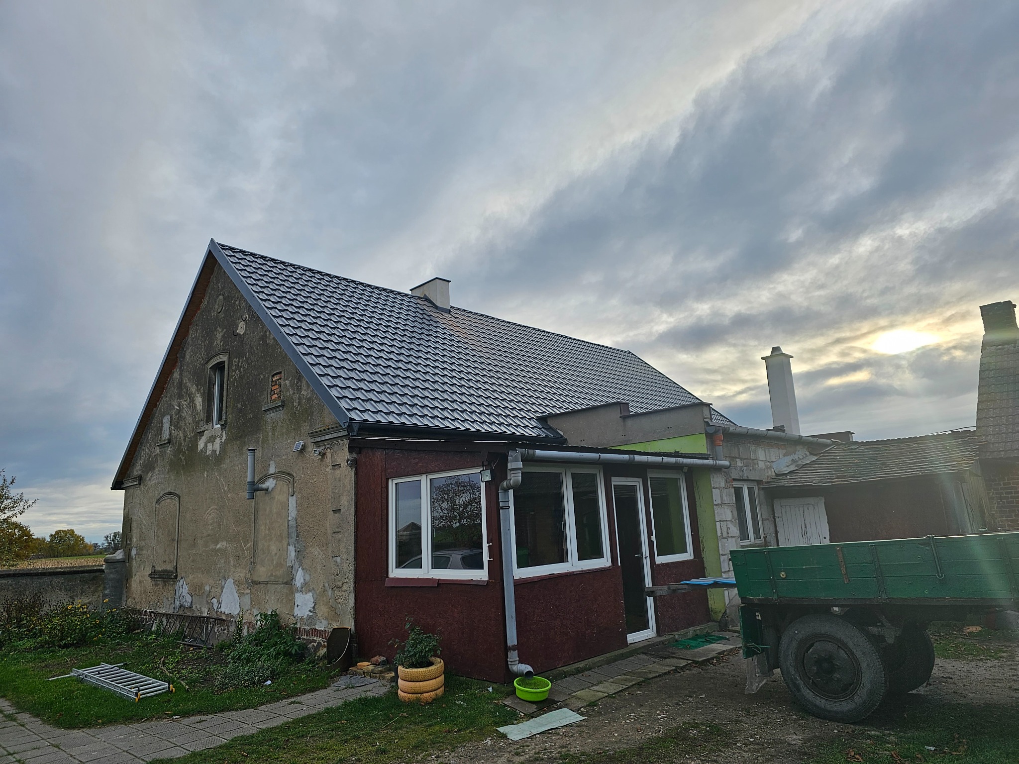 Dom z nowym, ciemnoszarym dachem z blachodachówki, widoczne rynny, stara elewacja, drabina oparta o ścianę i przyczepa rolnicza