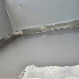 J.R.INVEST HOUSE JAN RATAJCZAK - Świeżo pomalowany narożnik balkonu z widocznymi metalowymi elementami i fakturą ściany w jasnym, szarym odcieniu.