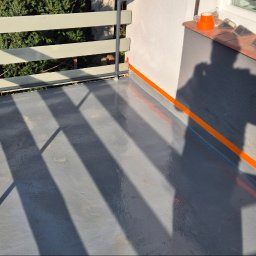 J.R.INVEST HOUSE JAN RATAJCZAK - Szary balkon z nową hydroizolacją, zabezpieczony pomarańczową taśmą malarską przy ścianie i balustradzie, cień osoby widoczny na podłodze.