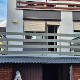 J.R.INVEST HOUSE JAN RATAJCZAK - Dwupiętrowy budynek z szarą elewacją, balkonem z drewnianą balustradą pomalowaną na szaro i roletami w oknach, z garażem w przyziemiu z blachodachówką i drabiną opartą o drzwi.