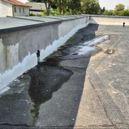 J.R.INVEST HOUSE JAN RATAJCZAK - Prace remontowe na dachu budynku: uszczelnianie ściany attyki i naprawa pokrycia z papy, widoczne ślady wilgoci i pracujący dekarz w tle.