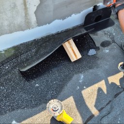 J.R.INVEST HOUSE JAN RATAJCZAK - Prace dekarskie: Obróbka blacharska dachu płaskiego z użyciem szlifierki kątowej i podkładki drewnianej, widoczne fragmenty uszczelnienia i odłamki materiału.