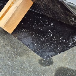 J.R.INVEST HOUSE JAN RATAJCZAK - Fragment balkonu z czarną folią hydroizolacyjną, drewnianym klockiem jako podpórką i resztkami białego cementu na szorstkiej, betonowej powierzchni.