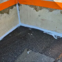 Spoina dylatacyjna wypełniona szarym uszczelniaczem na styku betonowej ściany i podłoża balkonu, zabezpieczona pomarańczową taśmą malarską.