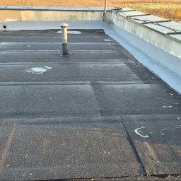 J.R.INVEST HOUSE JAN RATAJCZAK - Płaski dach pokryty papą z widocznymi kominkami wentylacyjnymi i nowo uszczelnionym obrzeżem, w tle pole.