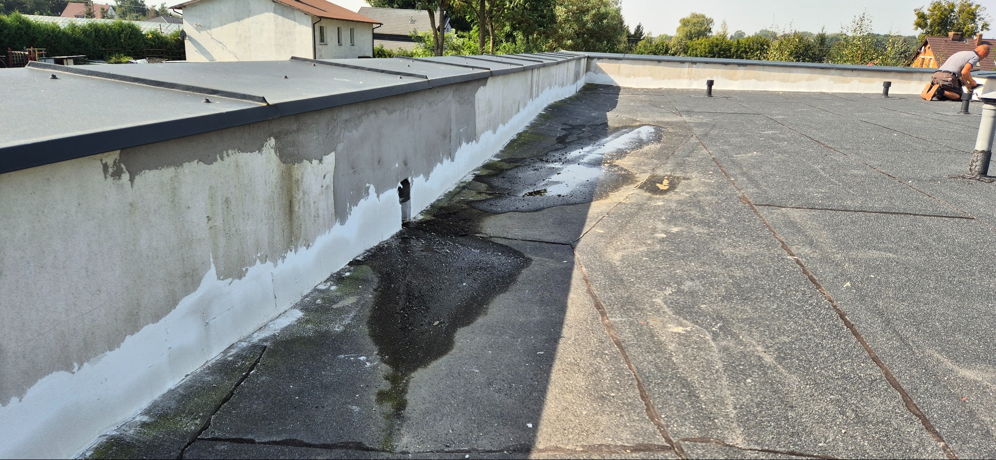 Prace remontowe na dachu budynku: uszczelnianie ściany attyki i naprawa pokrycia z papy, widoczne ślady wilgoci i pracujący dekarz w tle.