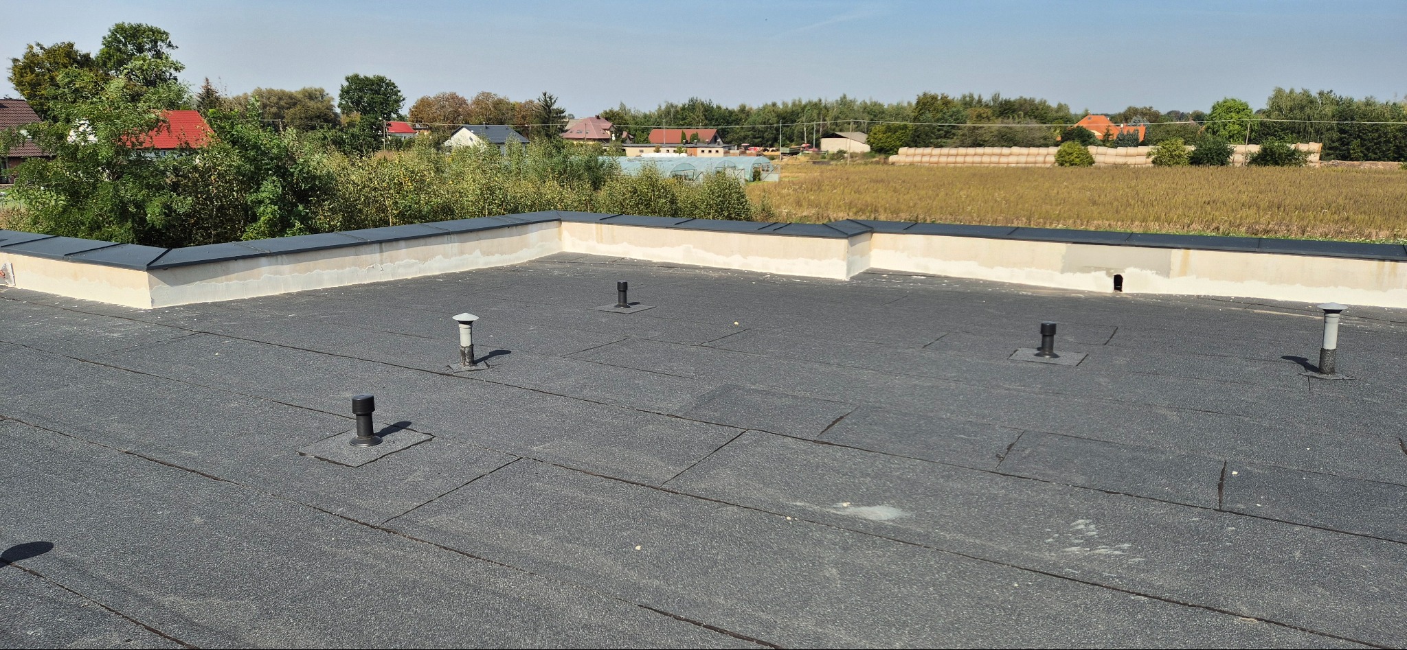 Widok z góry na płaski dach pokryty papą termozgrzewalną, z widocznymi kominkami wentylacyjnymi i obróbkami blacharskimi, w tle zabudowa jednorodzinna i pola uprawne.