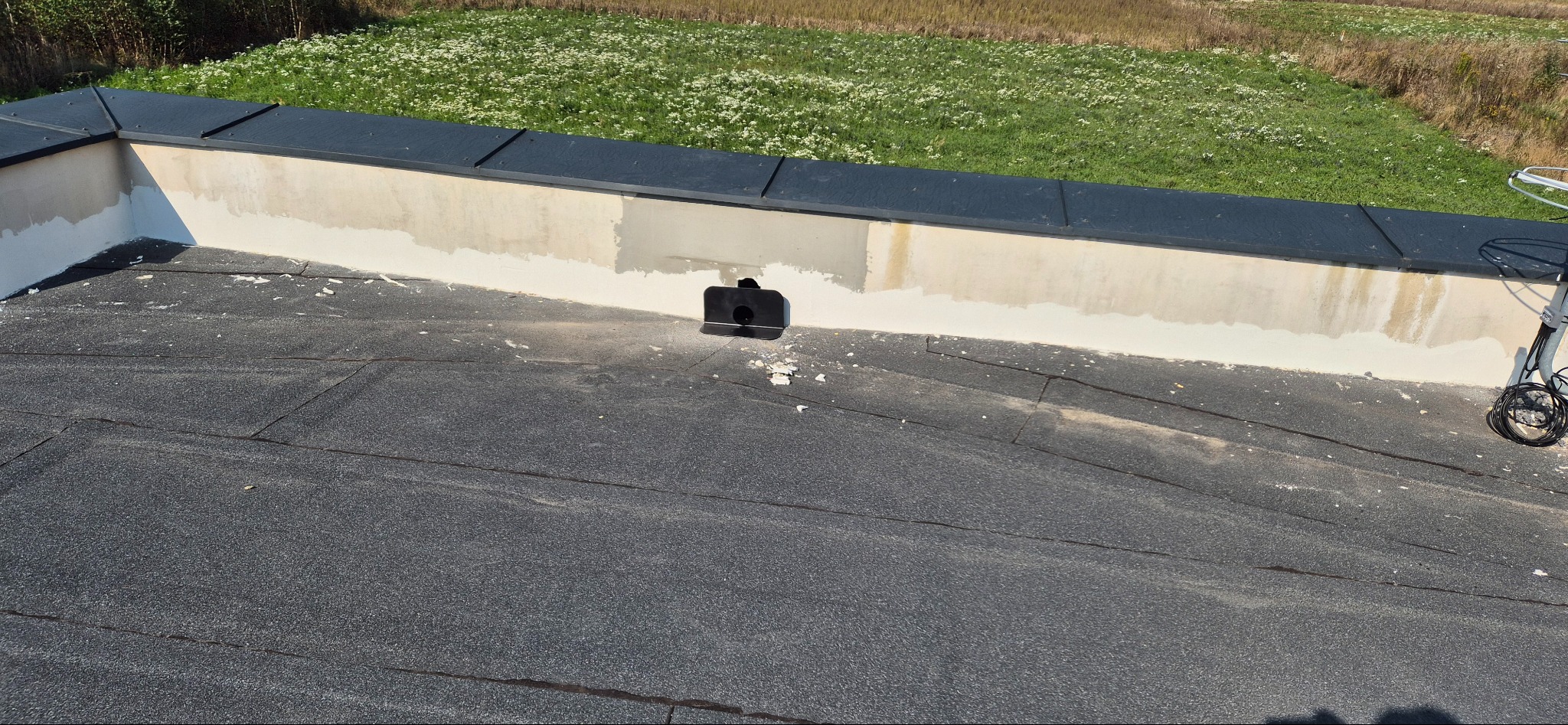 Widok z góry na fragment dachu pokrytego papą, z widoczną obróbką blacharską i fragmentem otynkowanej ściany attyki w trakcie prac wykończeniowych, z elementami świeżo nałożonej zaprawy...