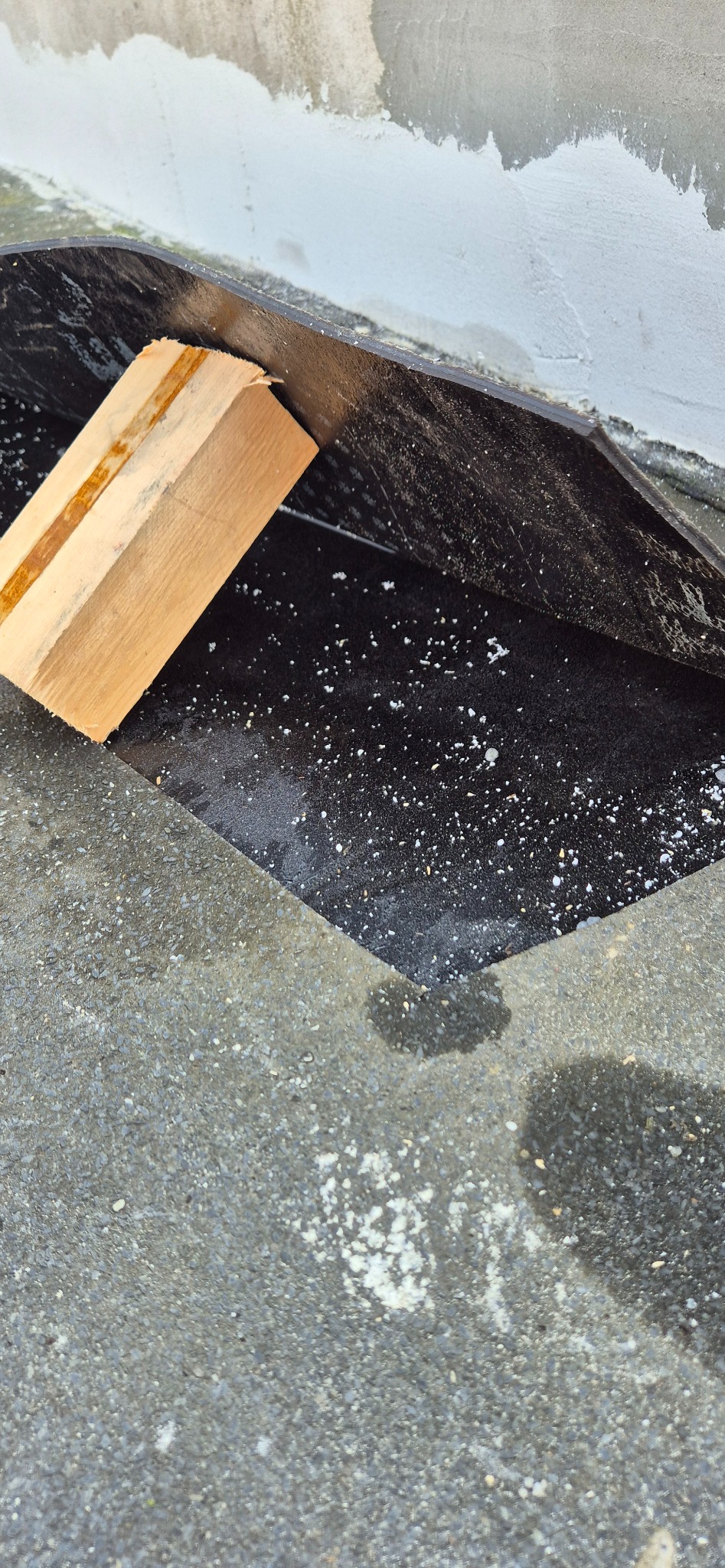 Fragment balkonu z czarną folią hydroizolacyjną, drewnianym klockiem jako podpórką i resztkami białego cementu na szorstkiej, betonowej powierzchni.