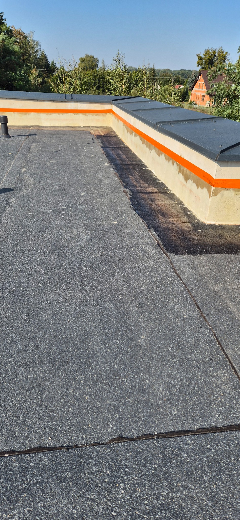 Widok z góry na płaski dach pokryty papą, z widocznymi detalami uszczelnienia na krawędziach, pomalowanymi na pomarańczowo, oraz kominkiem wentylacyjnym w tle.