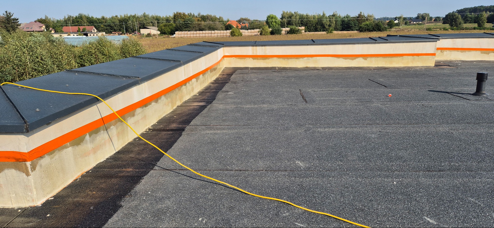 Wykończony płaski dach pokryty papą, z obróbkami blacharskimi i pomalowanym pasem na pomarańczowo przy attyce, widoczny przewód elektryczny.