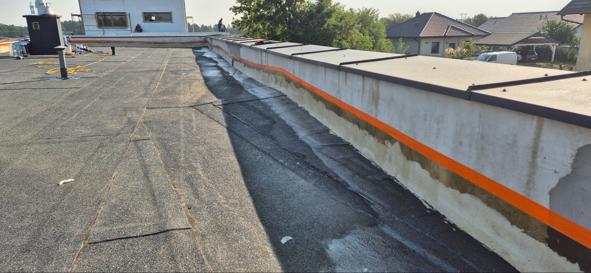 Wykończony dach płaski z papy bitumicznej z obróbkami blacharskimi przy attyce, widoczne ślady po niedawnym klejeniu i uszczelnianiu, w tle zabudowa jednorodzinna.