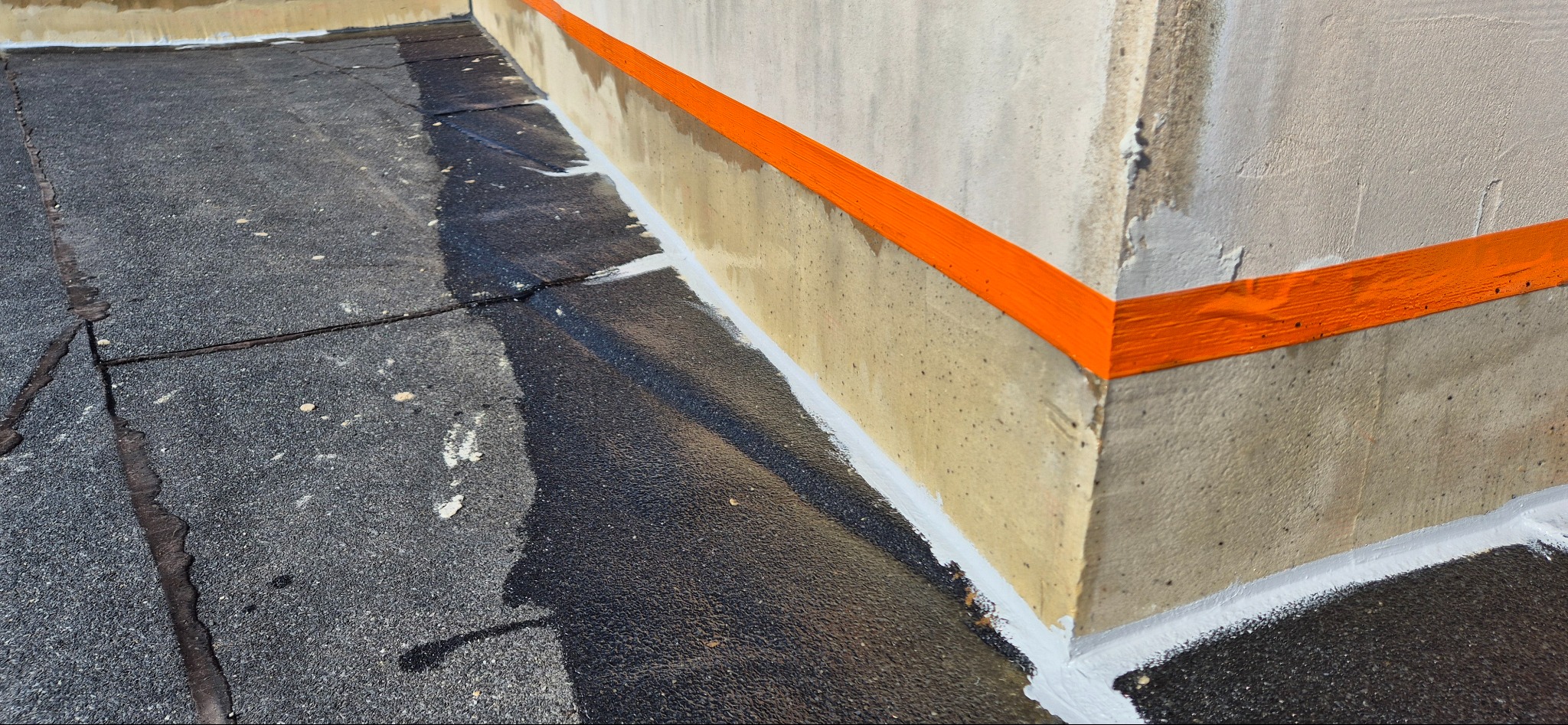 Szczegółowe ujęcie fragmentu dachu pokrytego papą, z widocznym uszczelnieniem przy ścianie oraz pomalowanym na pomarańczowo pasem na ścianie.