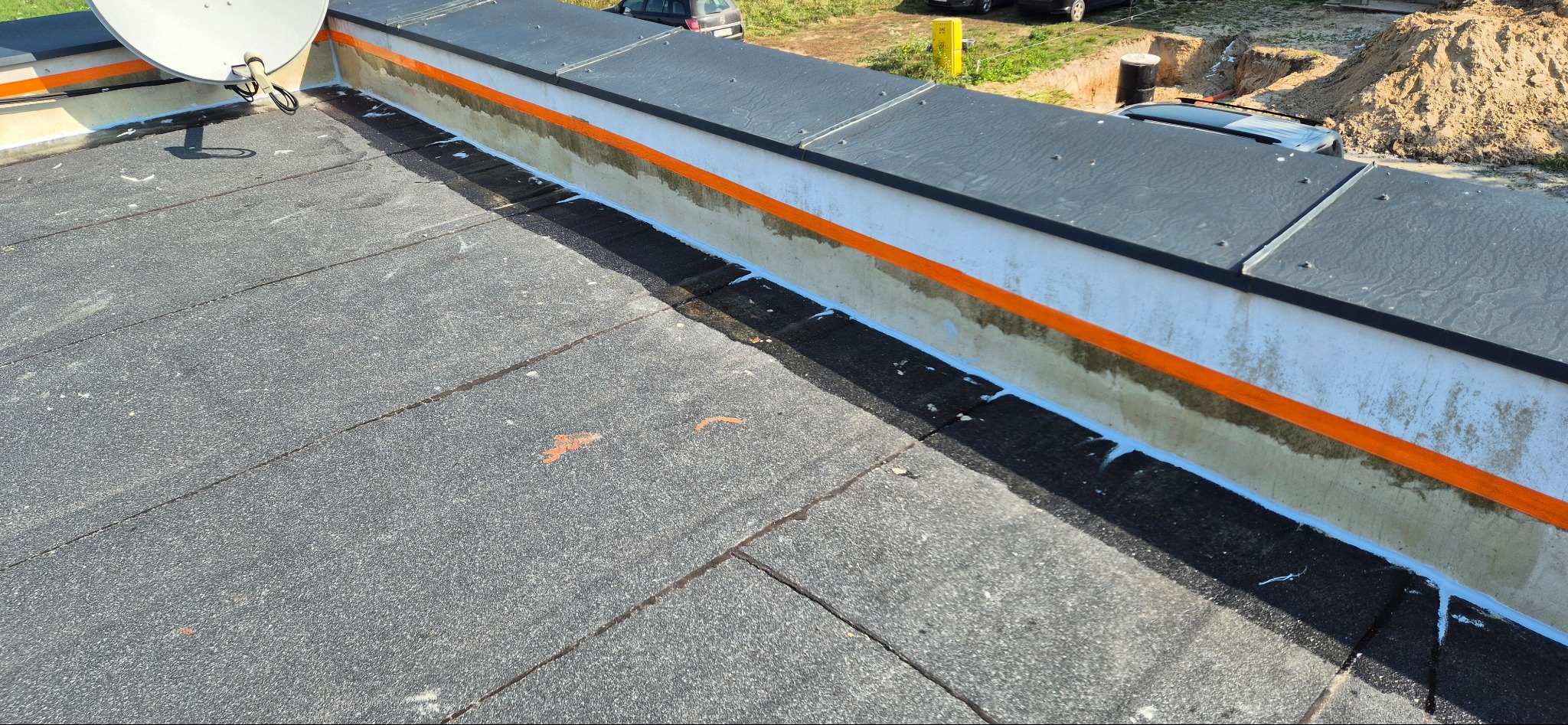 Wykończony fragment dachu płaskiego z papy, z widocznym obrzeżem zabezpieczonym pomarańczową taśmą i uszczelnionym białą masą, antena satelitarna w rogu.