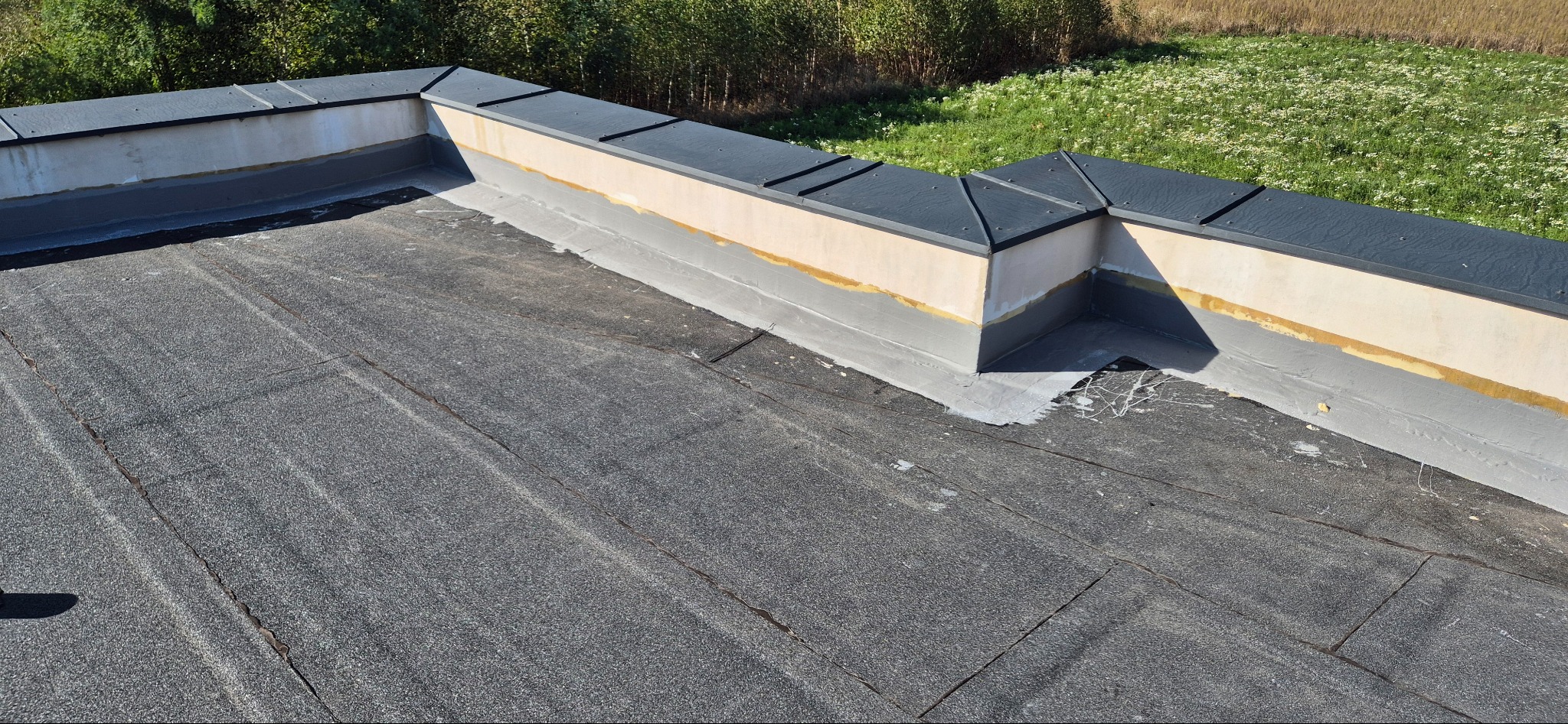 Widok z góry na płaski dach pokryty papą termozgrzewalną, z detalami obróbki blacharskiej i fragmentem attyki. Widoczne ślady po pracach dekarskich.