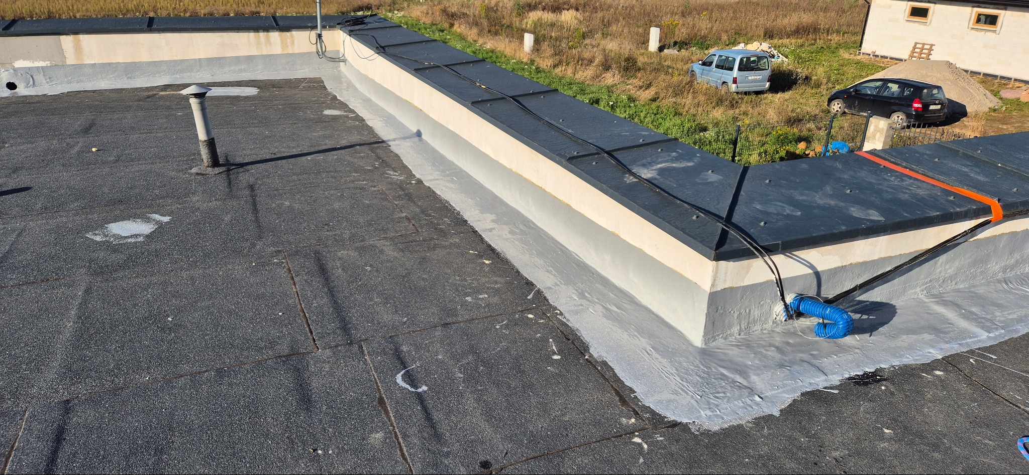 Widok z góry na płaski dach pokryty papą, z widocznymi obróbkami blacharskimi, kominkiem wentylacyjnym i elementami instalacji, w tle zaparkowane samochody i budynek.