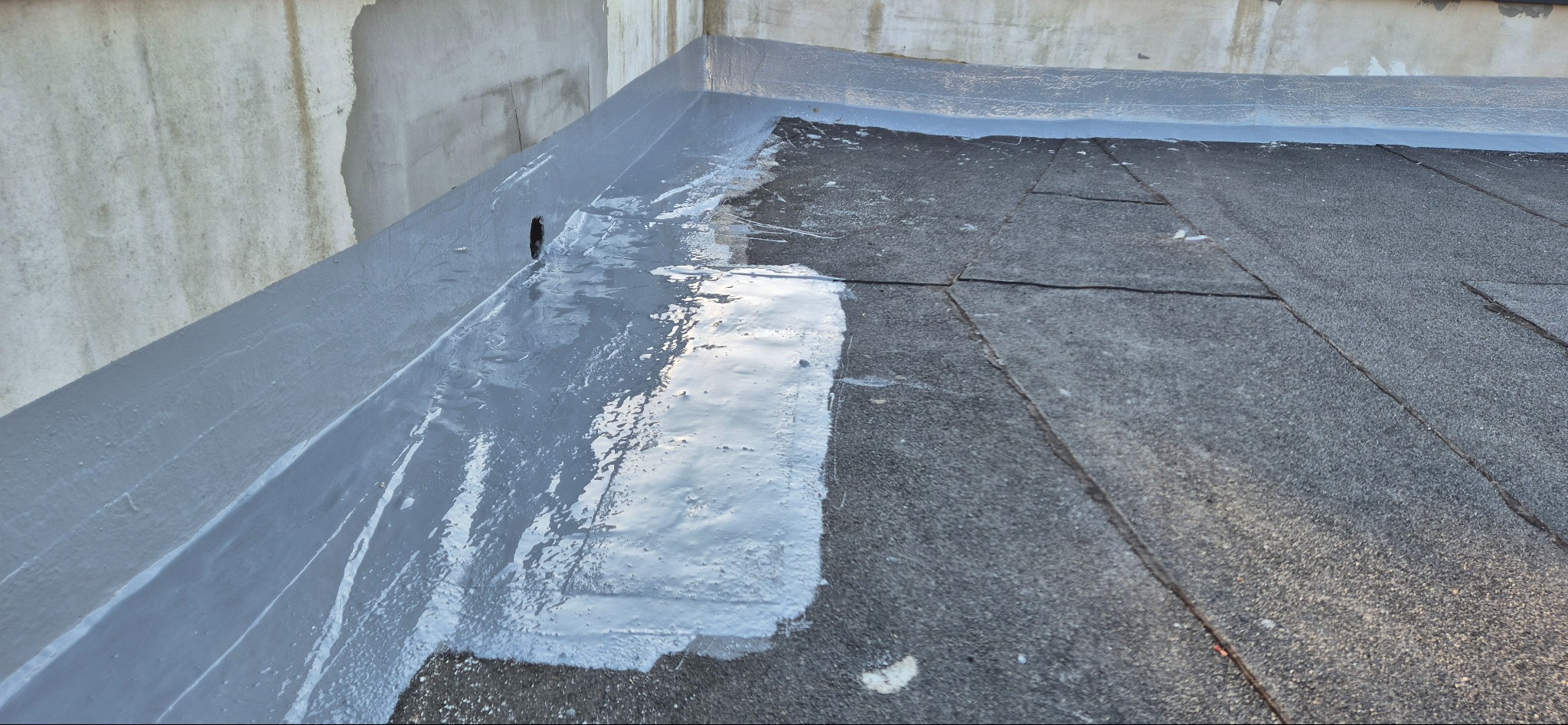 Świeżo uszczelniony, płaski dach pokryty papą, z widocznymi śladami szarej masy bitumicznej, szczególnie wzdłuż krawędzi i przy otworze wentylacyjnym, wskazując na niedawno wykonaną renowację...