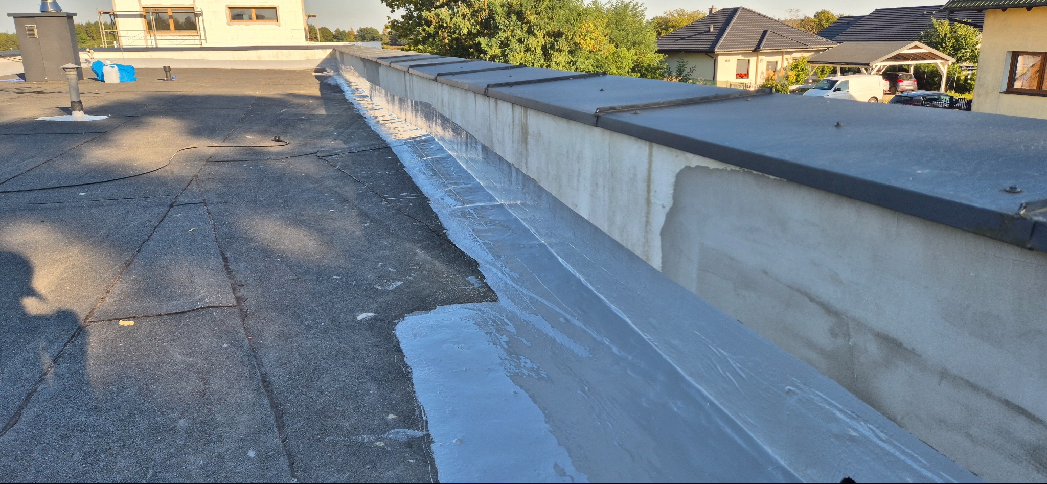 Płaski dach pokryty papą, z widocznym świeżo nałożonym szarym uszczelnieniem na łączeniu z attyką, w tle zabudowa jednorodzinna.