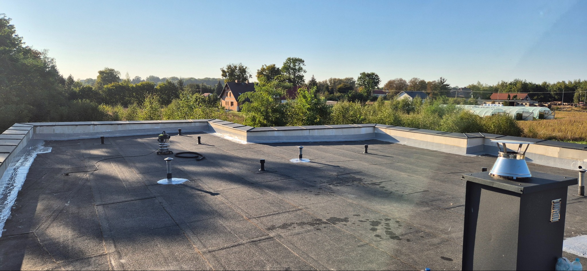 Widok z góry na płaski dach pokryty papą, z kominem wentylacyjnym i urządzeniem czyszczącym, w tle zabudowa jednorodzinna i zieleń.