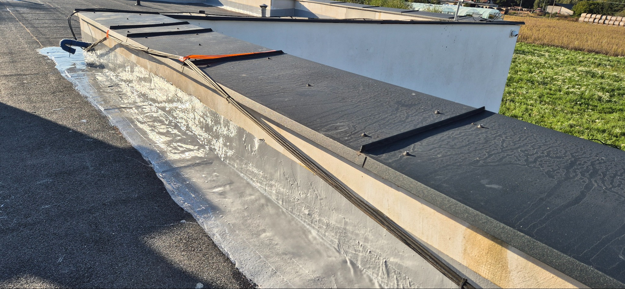 Wykończony fragment dachu płaskiego z widocznym zabezpieczeniem przeciwwodnym na krawędzi, z elementami mocującymi i przewodami na tle krajobrazu polnego.