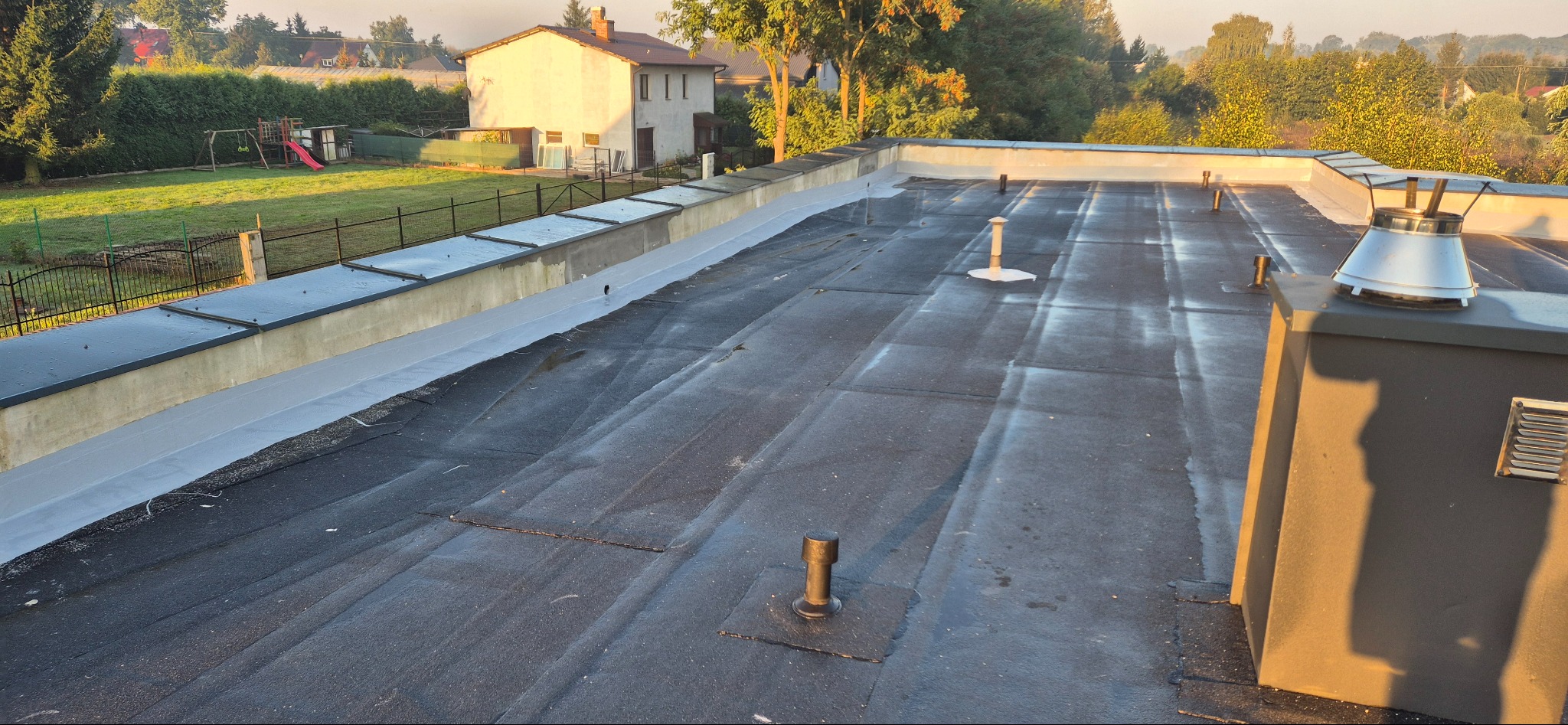 Widok z góry na płaski dach pokryty papą, z kominem i elementami wentylacyjnymi, w tle budynek mieszkalny i zieleń.