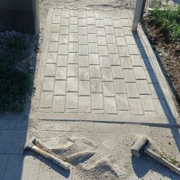 Yakiv Shevchenko - Układanie kostki brukowej na chodniku, widoczne narzędzia: młotek, przecinak, poziomica. Częściowo ułożona kostka, piasek, obrzeża. Prace brukarskie w toku.