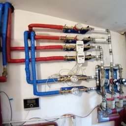 BUILD PROJECT SPÓŁKA Z OGRANICZONĄ ODPOWIEDZIALNOŚCIĄ - Rozbudowana instalacja wodna i grzewcza z rurami w izolacji termicznej (czerwonej i niebieskiej), liczniki wody, zawory, czujniki oraz panel sterowania na ścianie w pomieszczeniu technicznym.