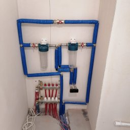 BUILD PROJECT SPÓŁKA Z OGRANICZONĄ ODPOWIEDZIALNOŚCIĄ - Instalacja hydrauliczna z filtrami wody i rozdzielaczem ogrzewania podłogowego, rury w niebieskiej izolacji, widok na surowy stan podłogi.